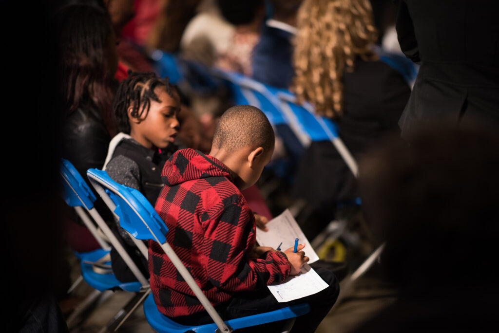 A photo of two young Black children, one boy and one girl, between the ages of 7 and 10. They are at a public town hall meeting, and are sitting next to each other on foldable chairs. Together they are filling out a paper survey.